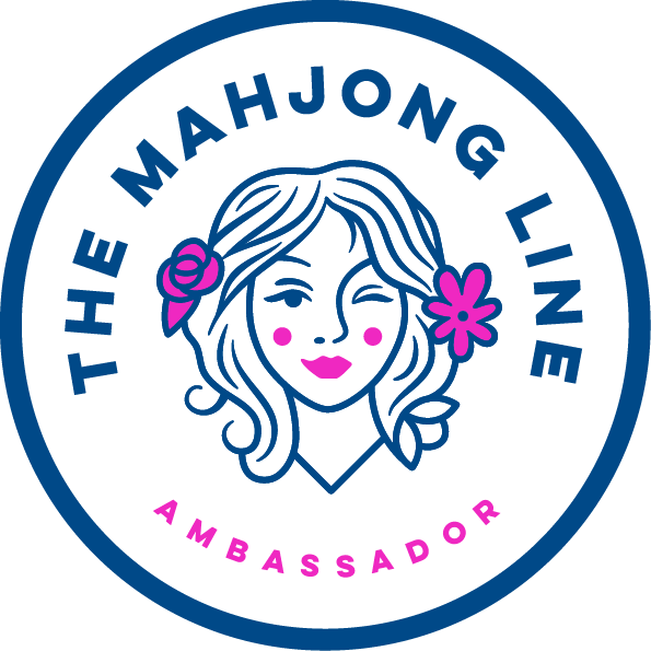 Logo for The Mah Jong Line featuring a cartoon woman with long wavy hair, pink flowers in her hair, and pink lipstick, with the words 'The Mah Jong Line' and 'Ambassador' surrounding her.