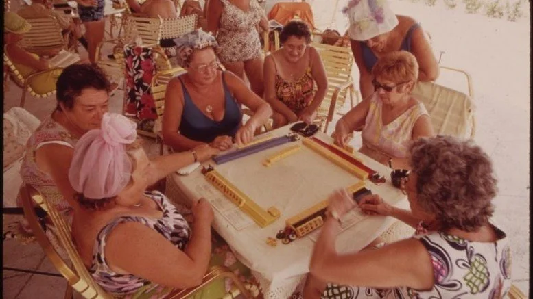 A group of women playing a game of mahjong at a table outdoors, with some women sitting and others standing nearby, in a casual social gathering.