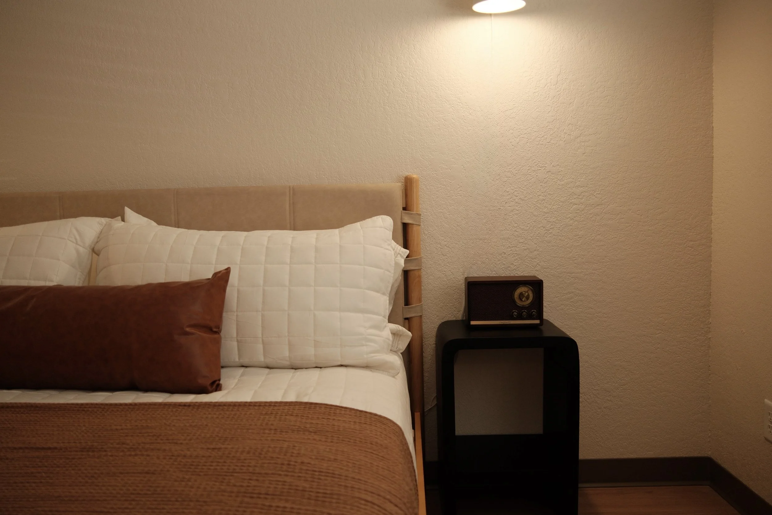 A bedroom with a bed featuring white quilted pillows and a brown pillow, a beige headboard, a black nightstand with a small vintage radio, and textured beige walls illuminated by a ceiling light.