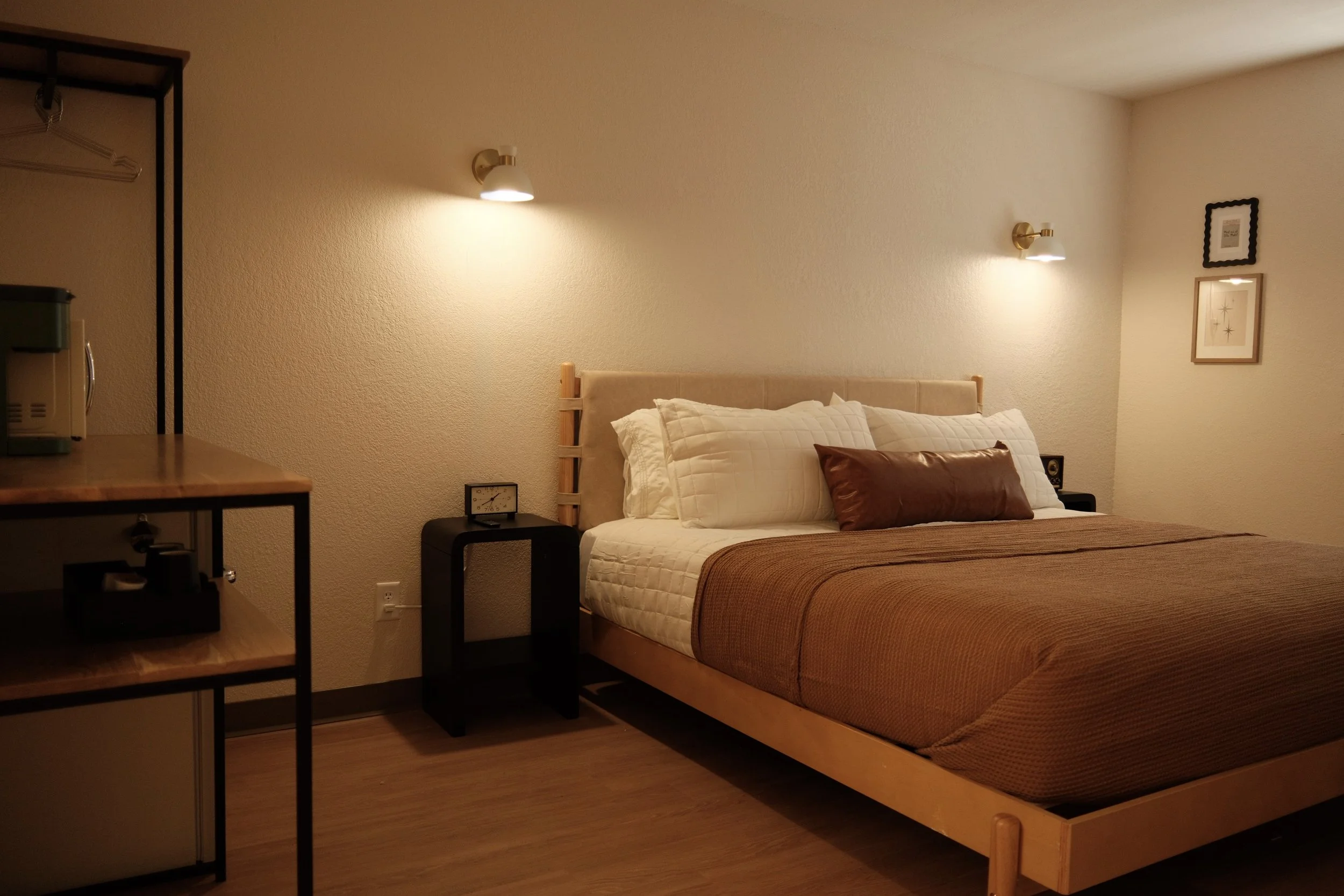A cozy bedroom with a double bed, brown bedspread, white pillows, and wall-mounted lamps above the bed. There are framed pictures on the wall, a small black nightstand with a clock, and a shelving unit on the left side.