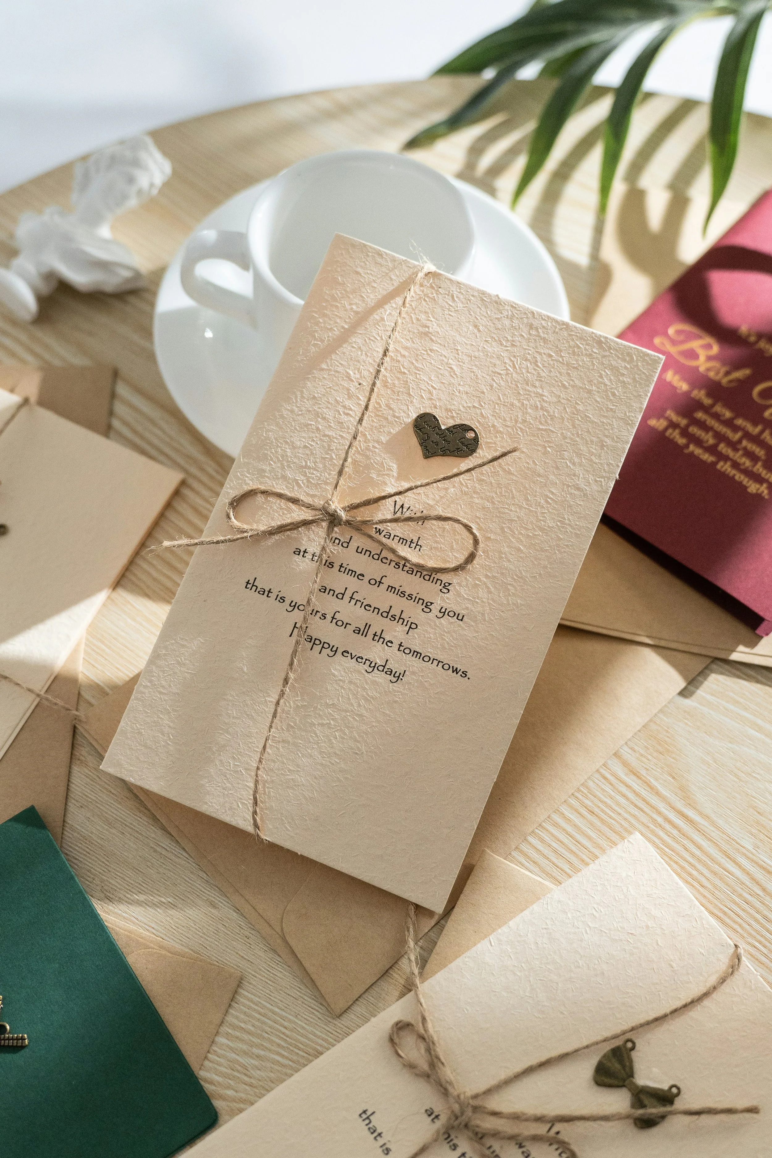 A beige textured card with a small jar lid heart sticker, tied with a piece of twine, holding a printed message, on a wooden surface with a white coffee cup and a green plant in the background.