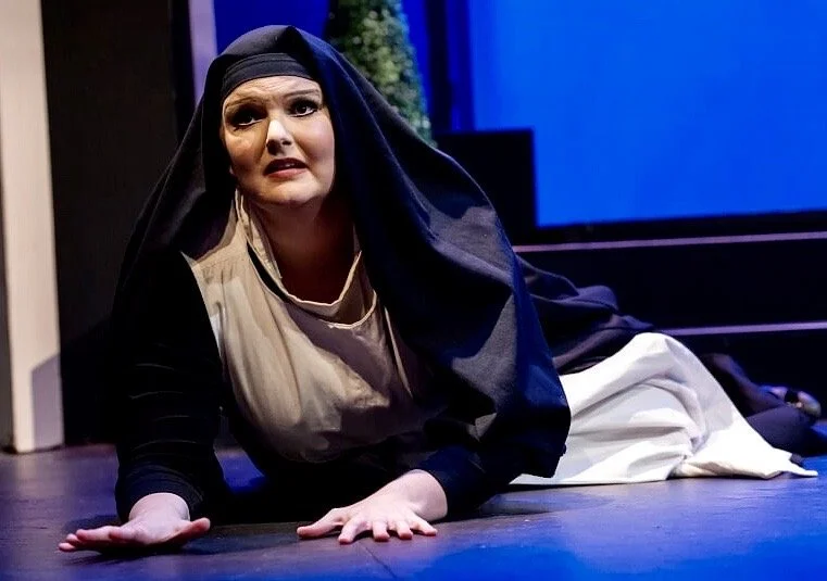 A woman dressed as a nun lying on the floor, looking distressed, on a stage with a dark background.