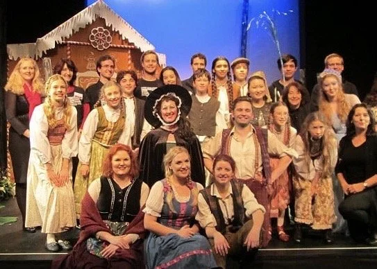 Group of people dressed in traditional costumes on stage, with a festive backdrop and a gingerbread house