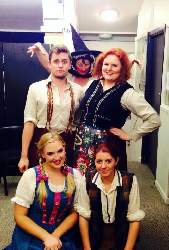 Five people in costume, with one person dressed as a witch with a purple hat and face paint, posing indoors with a black backdrop and white walls.