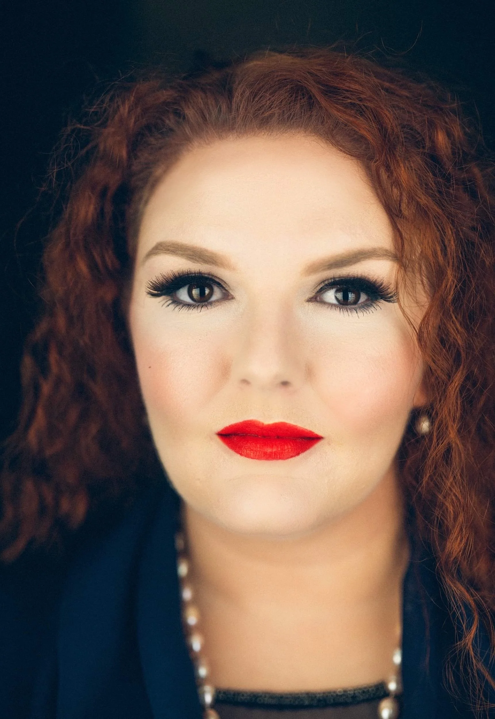 Portrait of a woman with curly red hair, red lipstick, and pearl earrings.