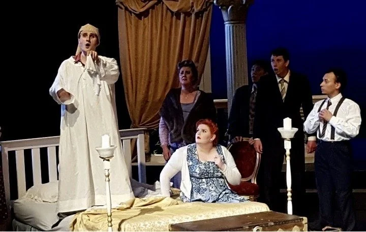 Group of six actors performing on stage, with one woman standing on bed, speaking or singing, and five others watching or listening, in a scene set with curtains and classical pillars.