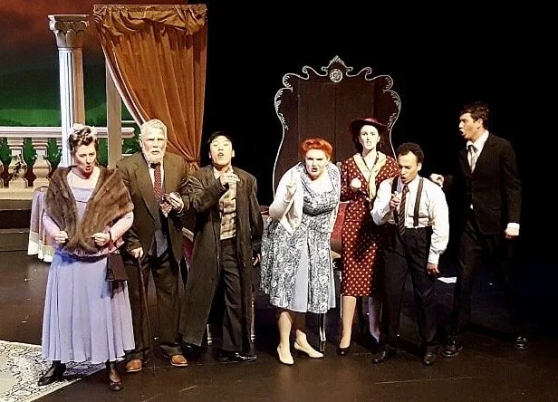 Stage scene with seven actors dressed in vintage clothing, standing in a line in front of a theater set with a curtain, columns, and a mirror, suggesting a theatrical performance.