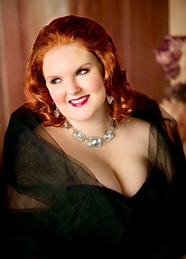 A woman with red hair wearing a black sheer top, a diamond necklace, and large earrings, smiling and looking to her right, with a blurred background.