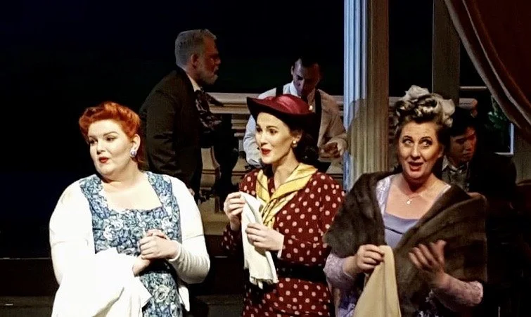 Three women in vintage 1950s clothing on a stage, with other people in the background, during a theatrical performance.