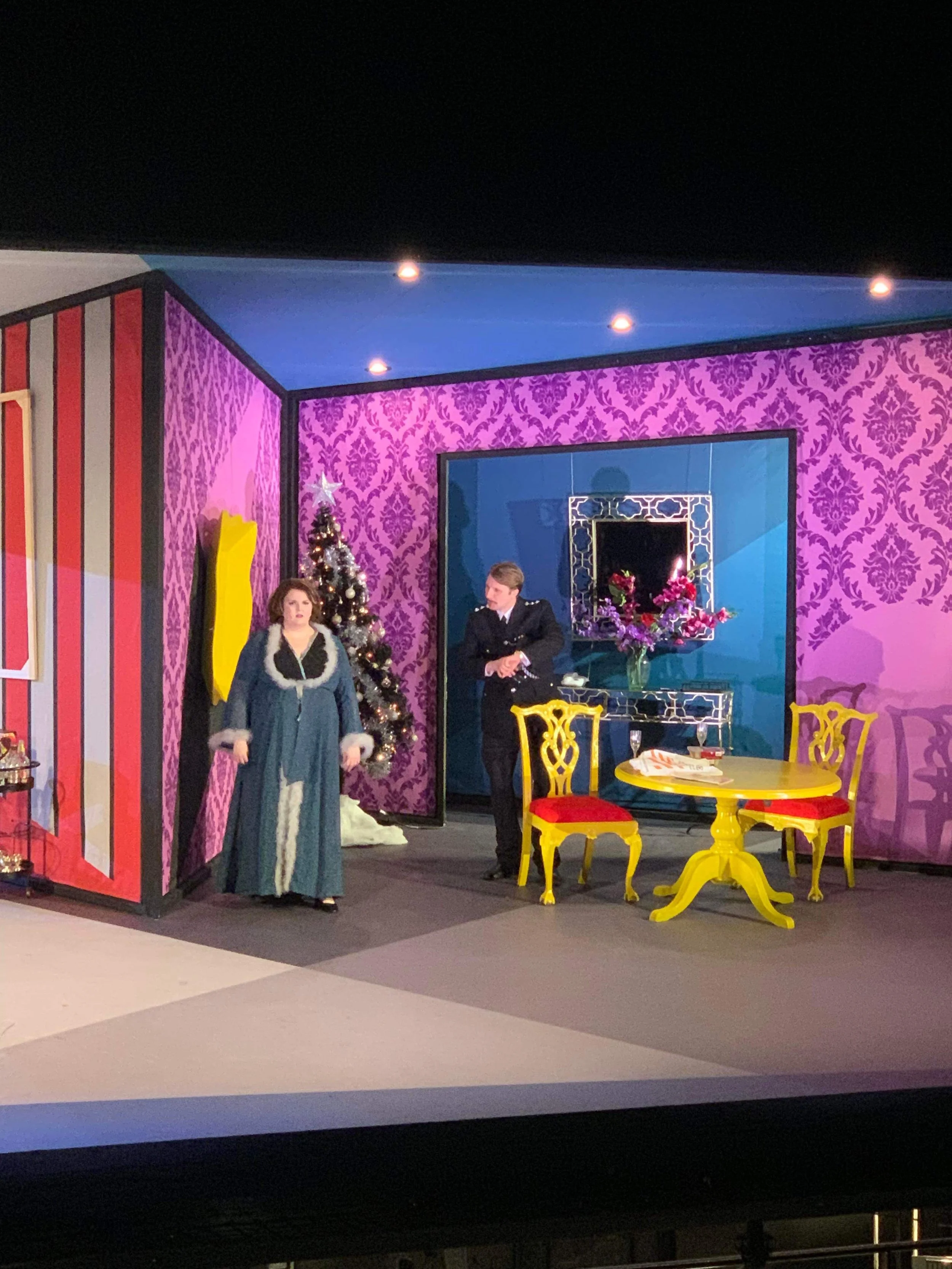 A theatrical stage set with a woman in a long blue dress and a man in a black suit, standing near a decorated Christmas tree, yellow and red chairs, and a table with flowers and a glass of wine, with vibrant pink, purple, and blue wallpapered walls.