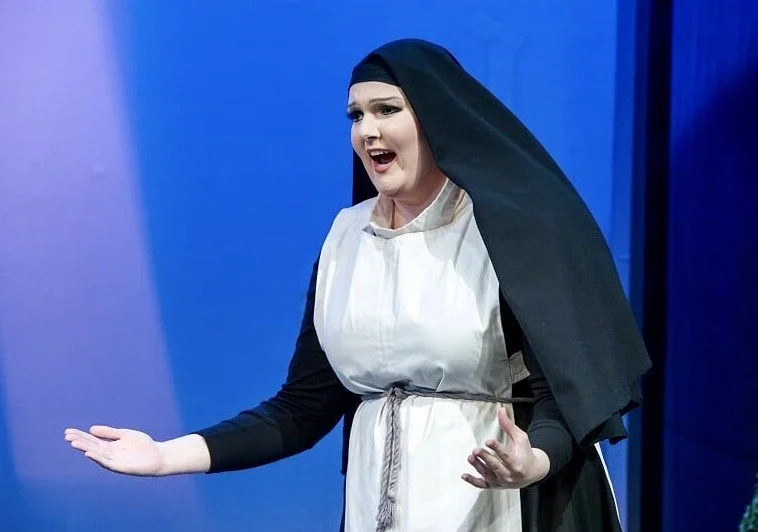 Woman dressed as a nun with a black veil and white dress, performing on stage against a blue background.
