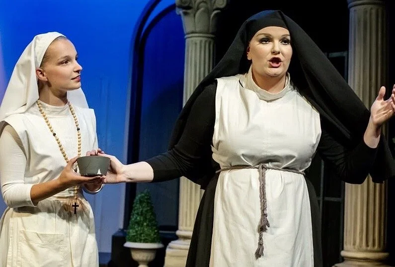 Two women in religious costumes performing on stage, one dressed as a nun and the other as a nun or religious figure, with a backdrop of classical columns and a potted plant.