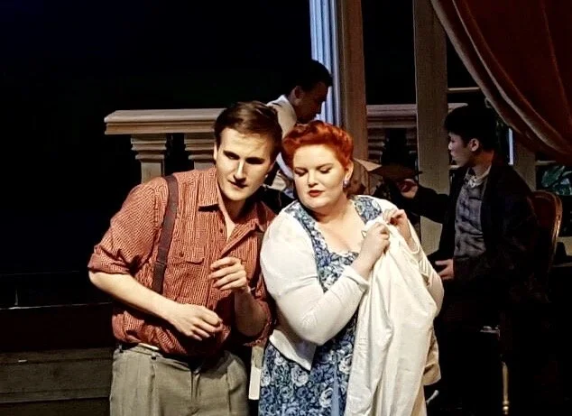 Three actors on stage performing in a theater, with two men and a woman in the foreground and a man in the background, engaged in a scene.