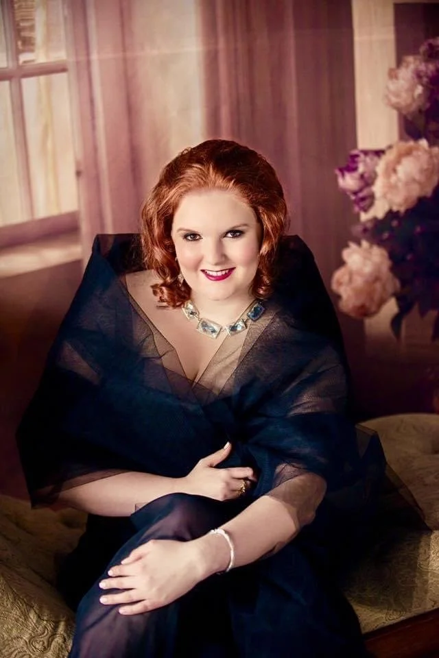 A woman with red hair, wearing a dark dress and a statement necklace, sitting on a sofa in a warmly lit room with flowers in the background.