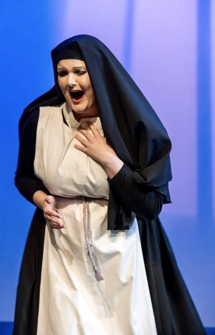 A woman dressed as a nun appears to be auditioning for a religious role, holding her chest and stomach with a distressed expression, in front of a blue background.