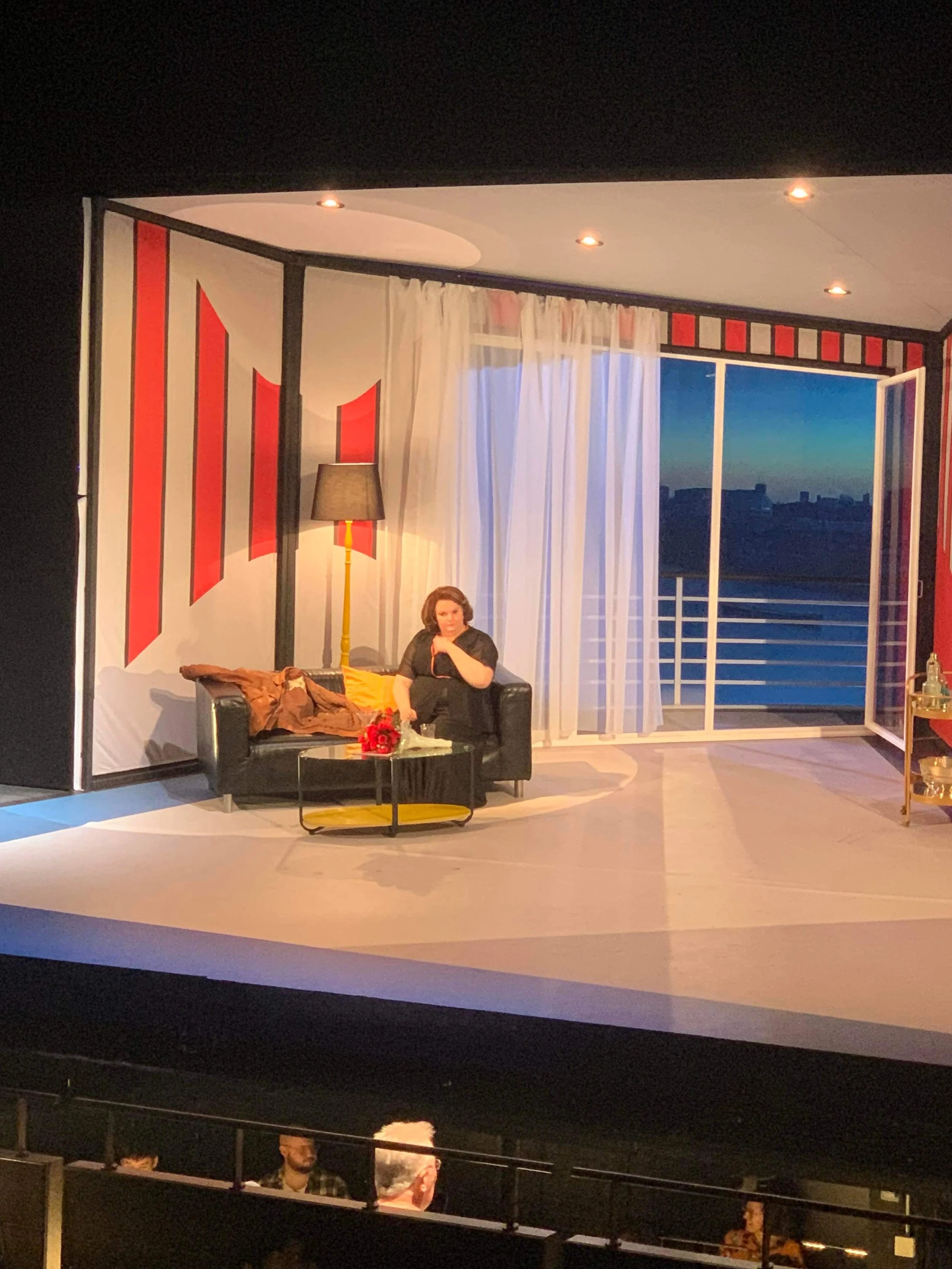 Actress sitting on a black couch on a theater stage set resembling a living room, with a window showing a blue sunset sky and city skyline in the background, white curtains, yellow cushions, a black coffee table with flowers and a cup, a tall floor l