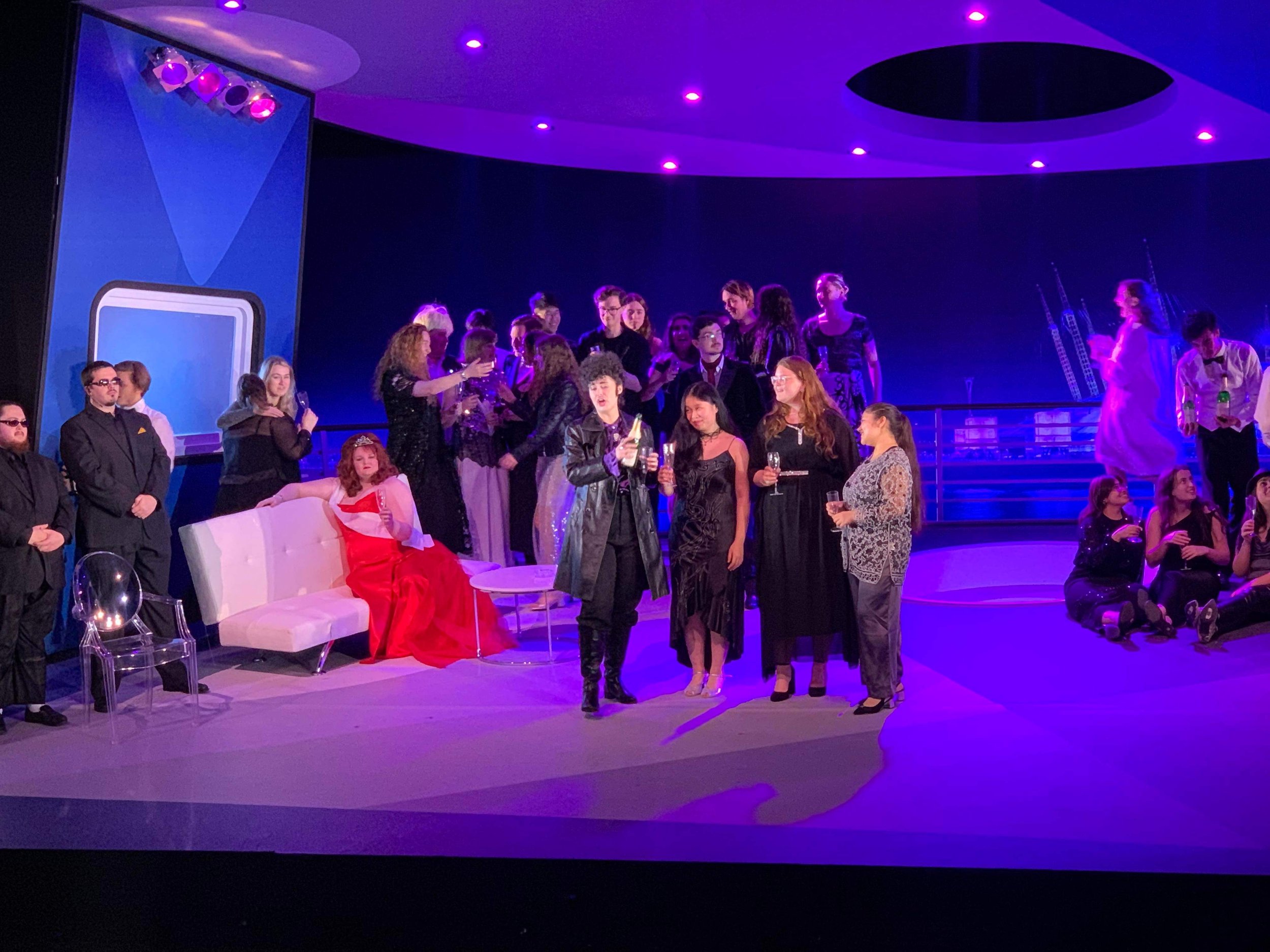 People at a stage party, with some sitting on a white bench, others standing and talking, in a dimly lit room with purple lighting and a large screen in the background.