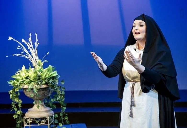 A woman dressed in historical or theatrical costume, wearing a black cloak and a beige dress with a rope belt, standing on a stage with a blue backdrop. She appears to be performing or speaking, with a floral arrangement nearby.