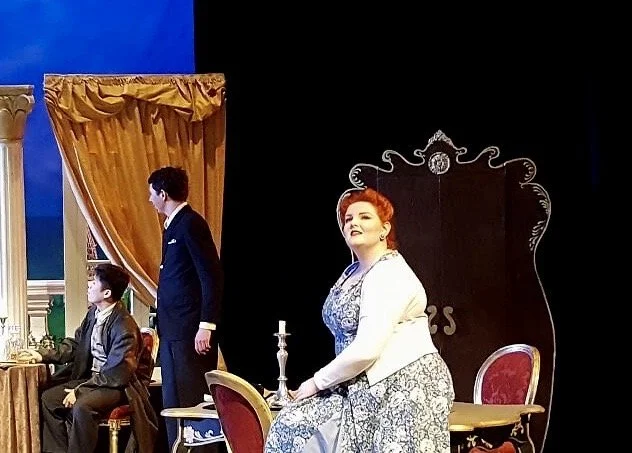 The image shows three people on a stage set for a play or performance. A woman with red hair, wearing a white jacket over a blue patterned dress, is sitting at a round table with a red upholstered chair and a candelabra. Two male actors are standing 