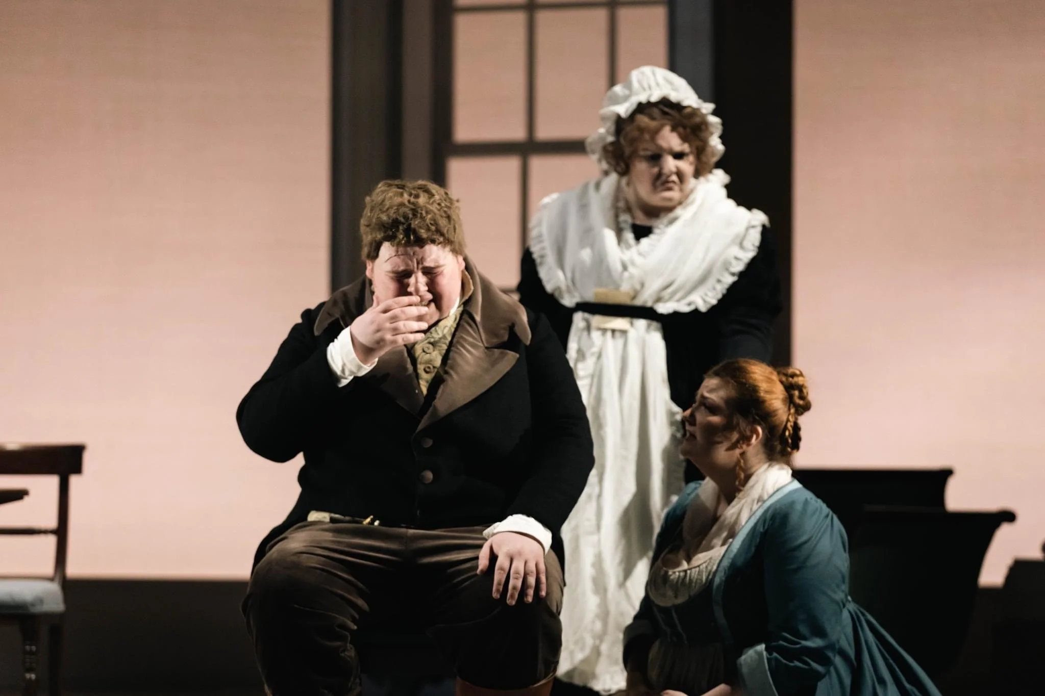 Three actors in period costumes performing on stage; one man appears to be crying or upset, a woman kneels nearby, and another woman stands behind them with a stern expression.
