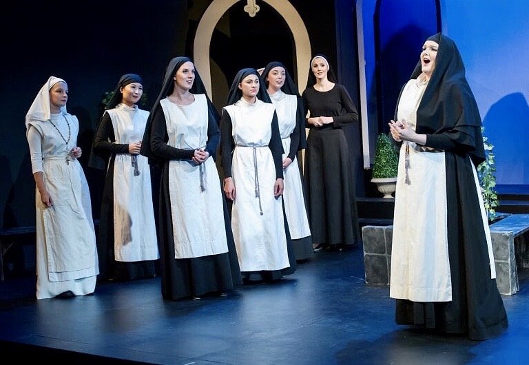 A theatrical performance on stage depicting nuns, with five women dressed as nuns and one woman in a black dress, engaged in a scene.