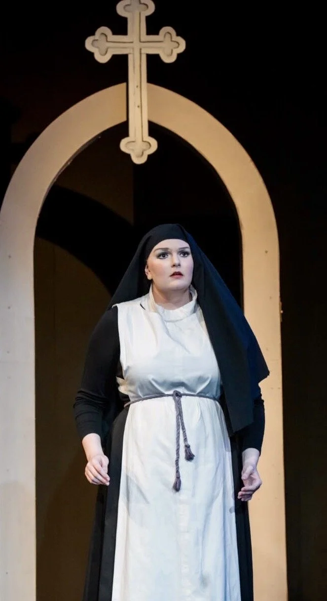 A woman dressed as a nun standing on stage with a religious cross above her head.