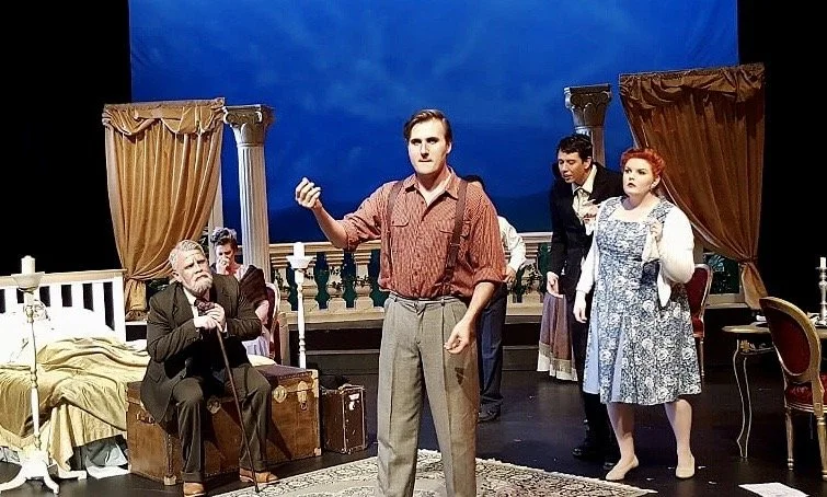 Cast performing a scene on stage, with actors in period costumes, a set resembling a bedroom and balcony, and a blue sky backdrop.