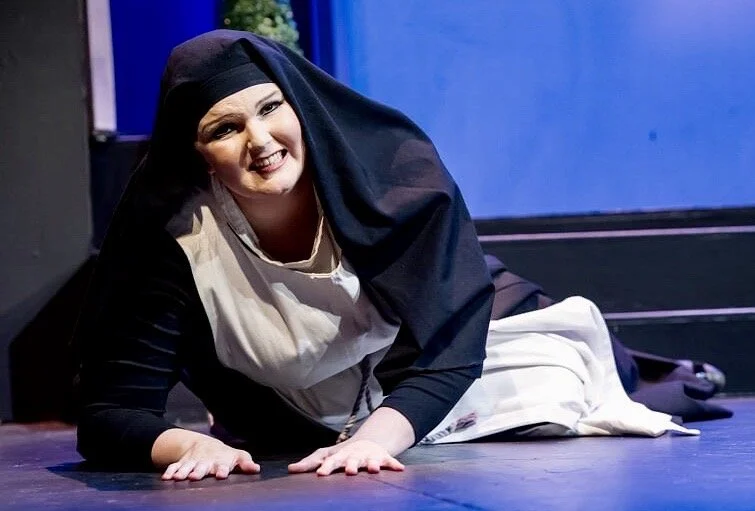 A woman dressed as a nun lying on the floor, smiling at the camera, with a black and white habit and a dark blue background.