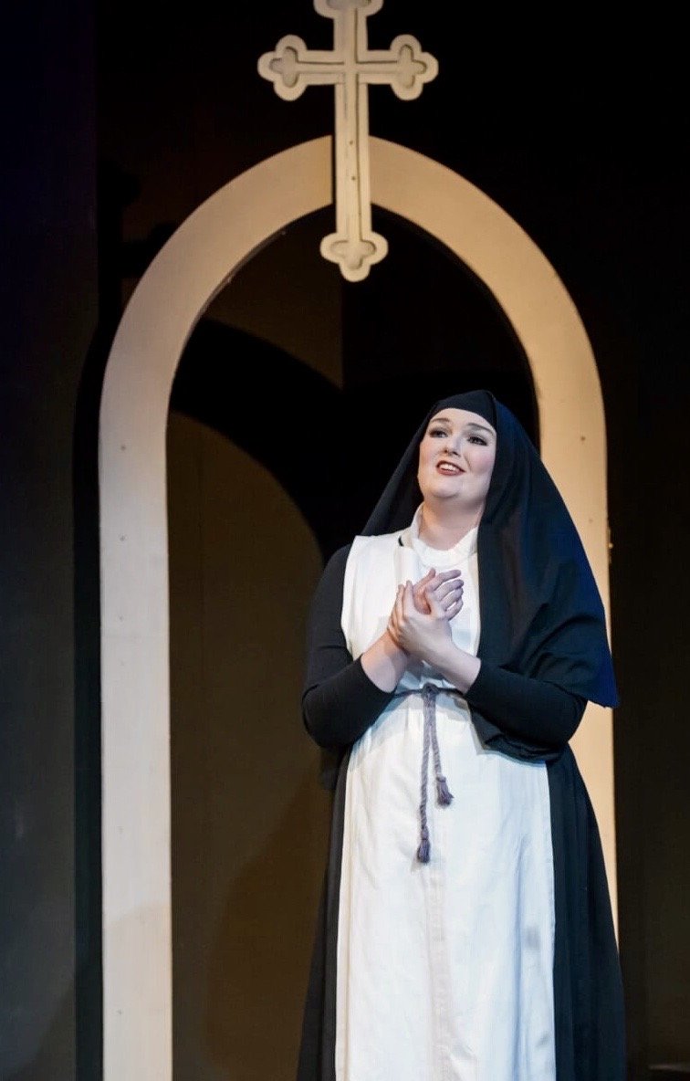 A woman dressed as a nun stands on stage in front of a religious backdrop with a cross and archway, performing with her hands clasped.