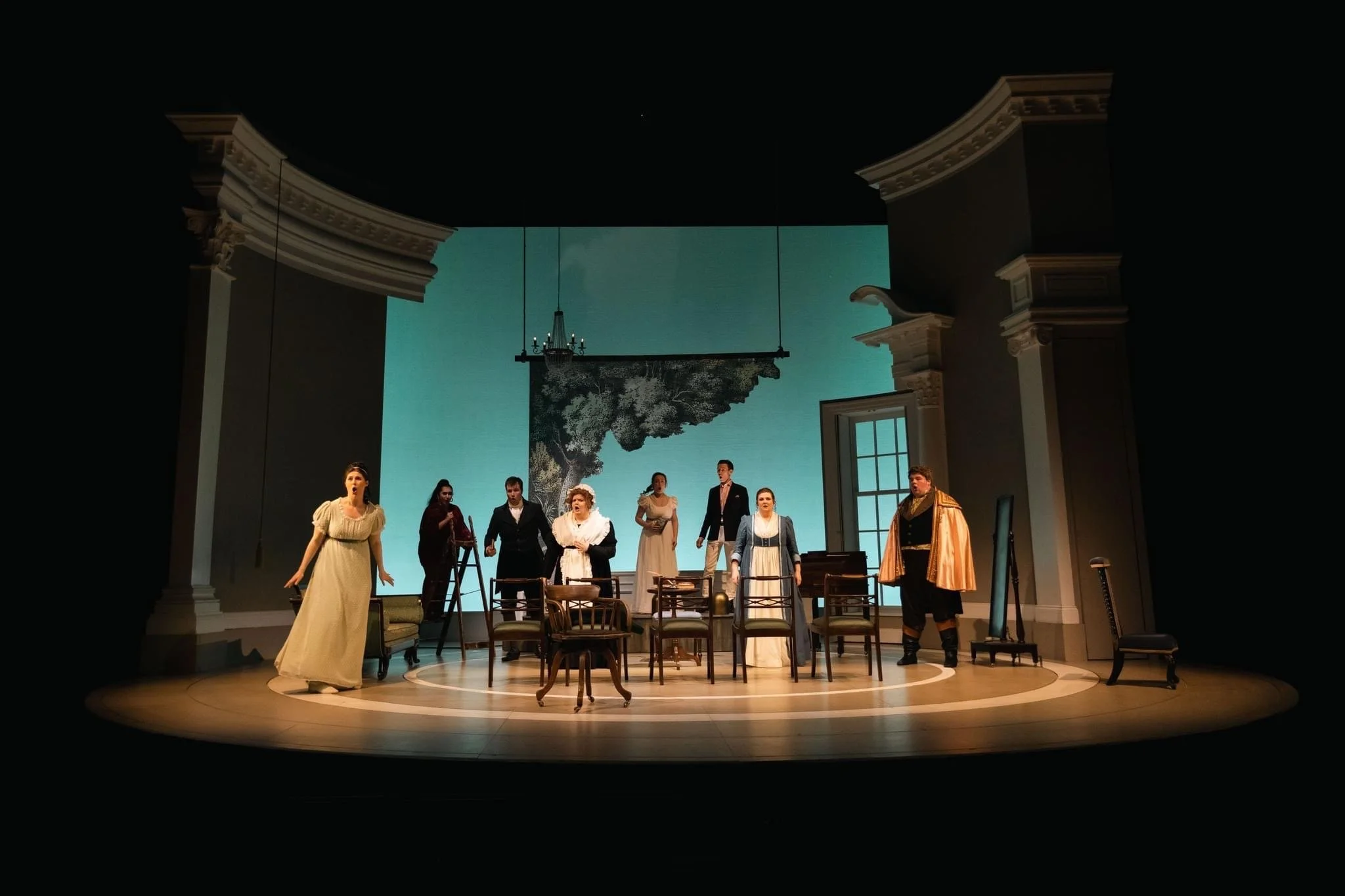 Theater stage with actors performing a scene in period costumes, set in an interior with a large backdrop showing a tree and a window, surrounded by decorative columns.