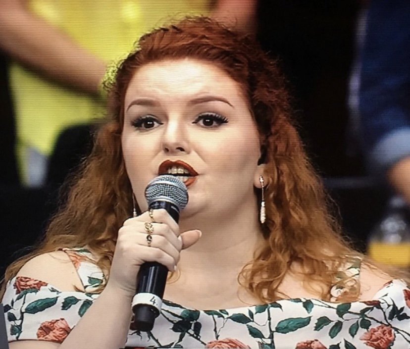 A woman with red curly hair holding a microphone and speaking.