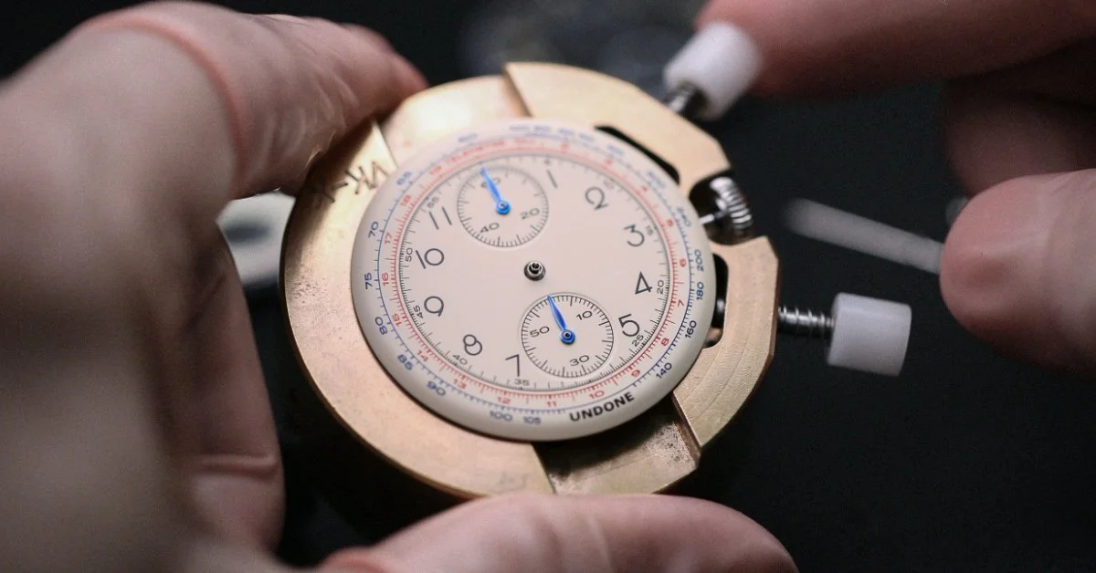 A watchmaker's hand working on a vintage chronograph watch movement, with tools gripping the movement for assembly or repair.