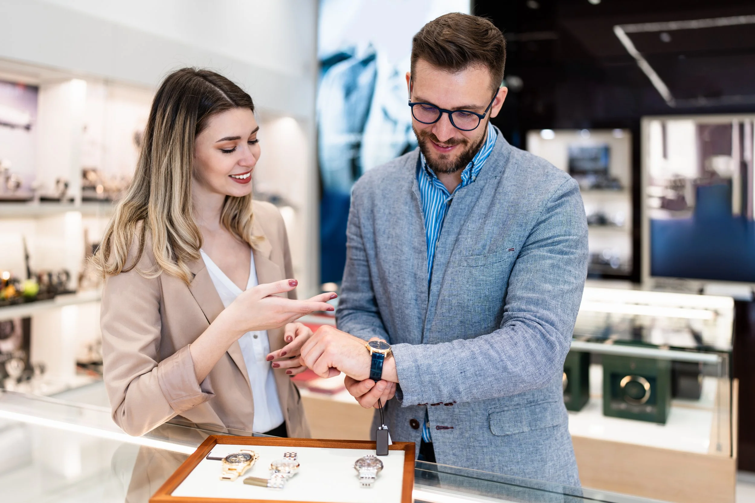 A man and woman shopping for watches in a store, with the man trying on a watch while the woman points at it and smiles.