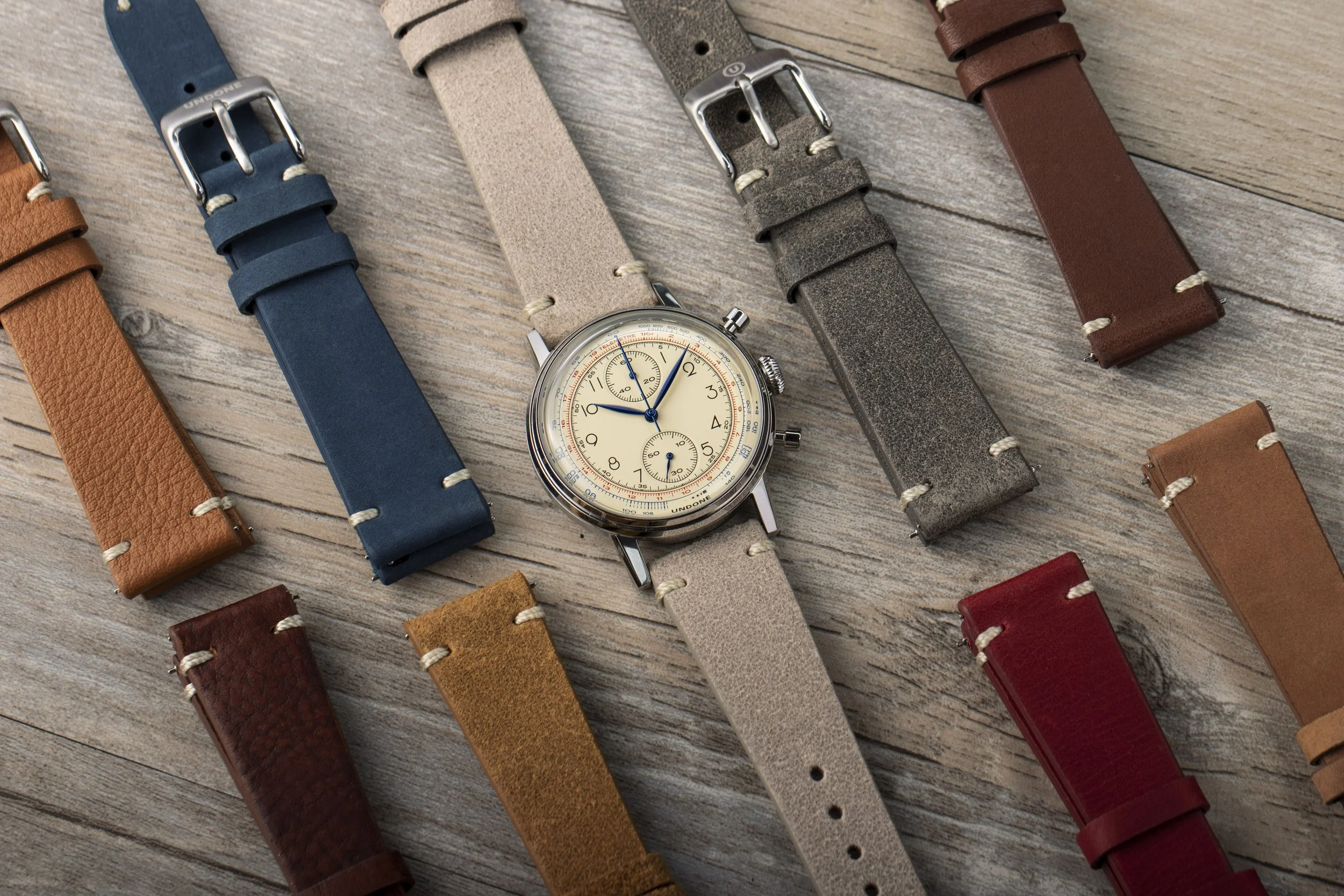 A variety of leather watch straps in different colors arranged around a vintage-style chronograph watch on a wooden surface.
