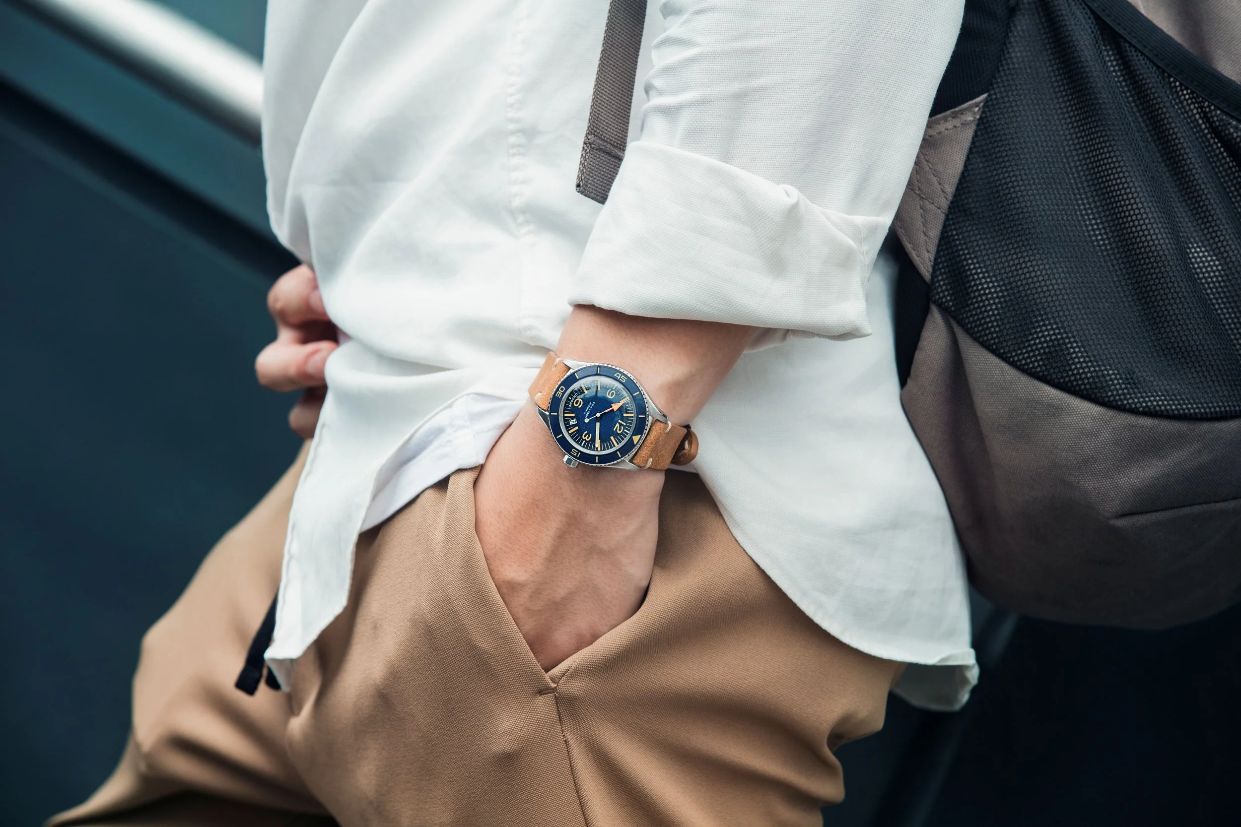 Person wearing a beige watch on their left wrist, dressed in a white long-sleeve shirt with sleeves rolled up and khaki pants, standing outdoors with an arm crossed.