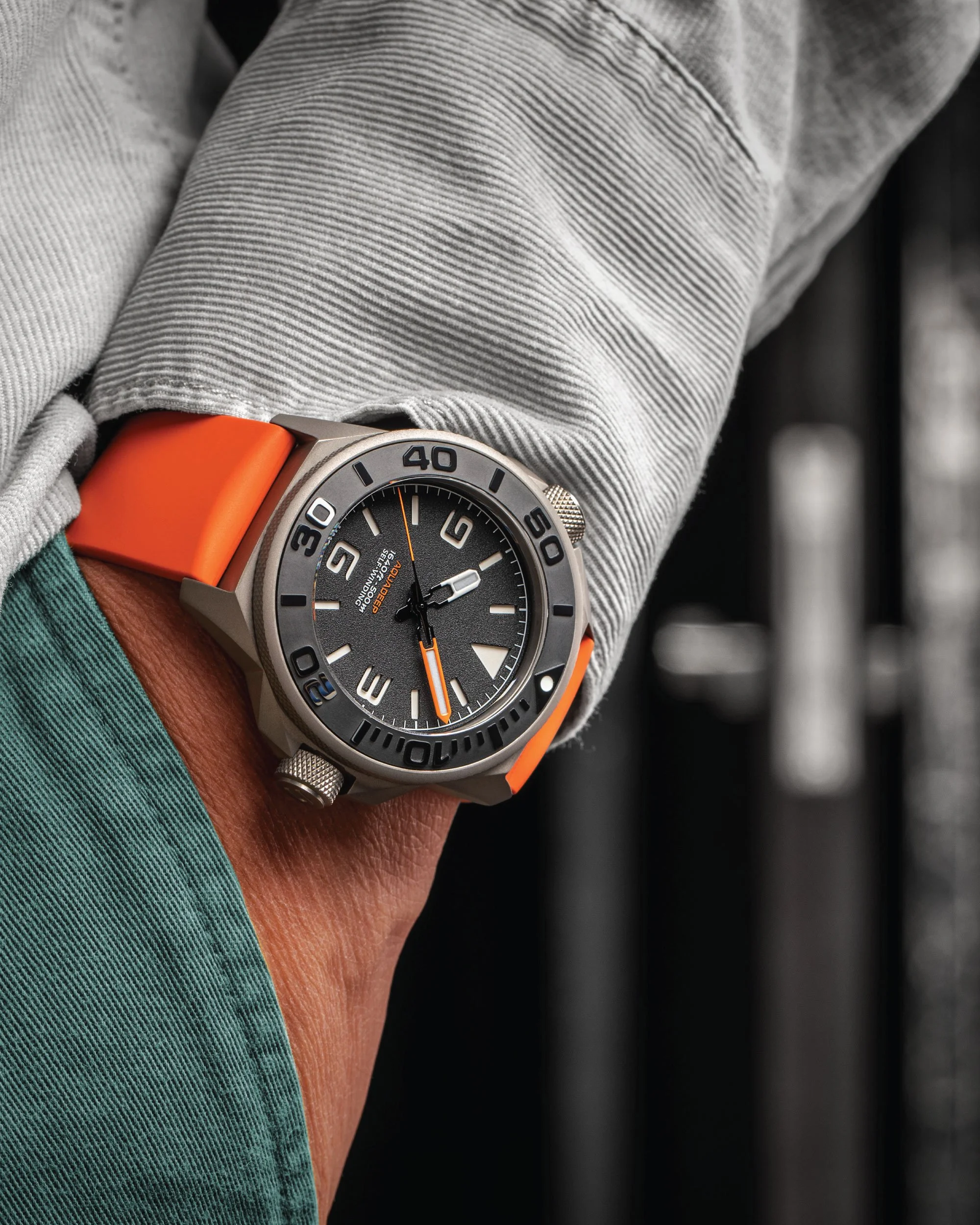 Close-up of a person's wrist wearing a wristwatch with an orange strap, against a background of a gray and green fabric, possibly clothing.