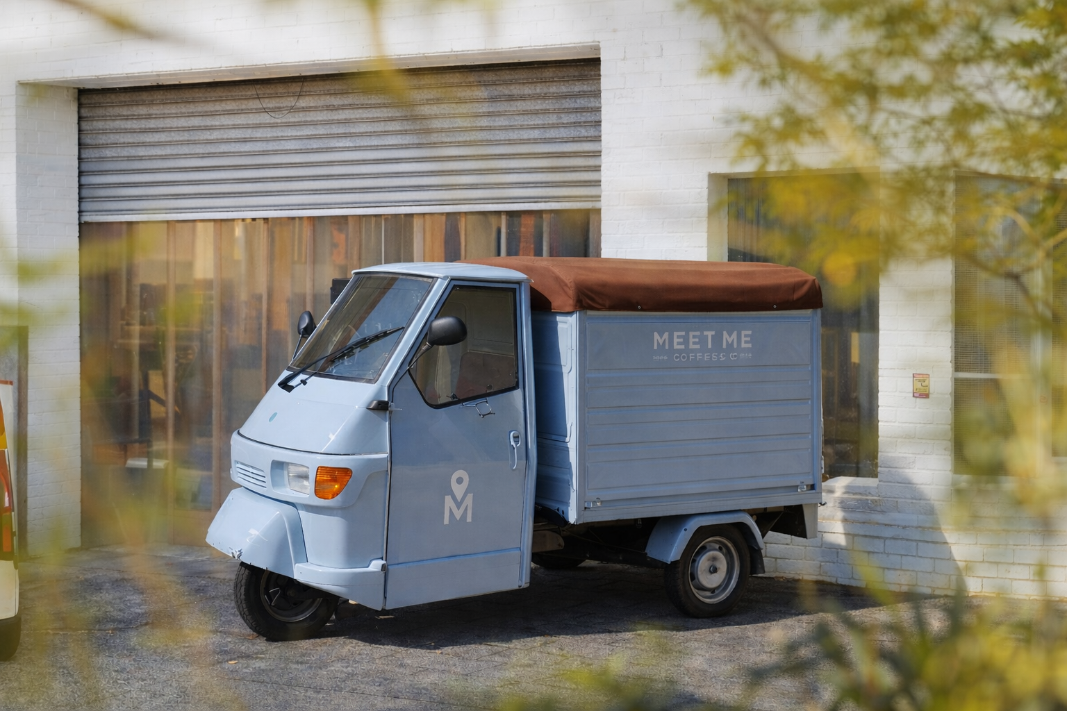 A light blue three-wheeled delivery vehicle with a brown canopy, parked in front of a white brick building with a garage door and a window, with yellow leaves partially obscuring the view.