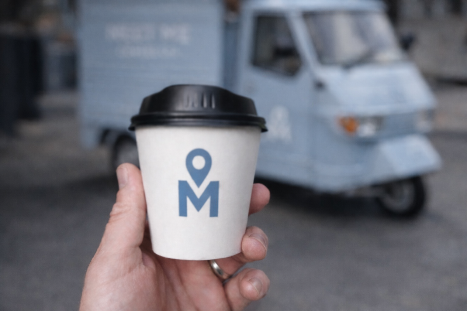 Hand holding a disposable coffee cup with a black lid and a logo featuring a map pin and the letter M, with a blurred street scene and a small vehicle in the background.