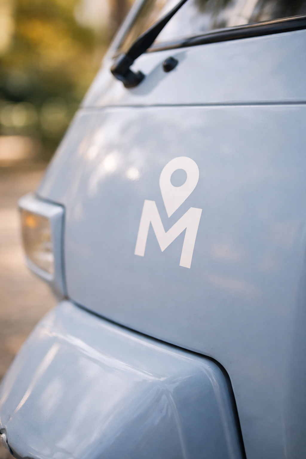 Close-up of the rear side of a light blue vehicle with a white logo featuring a location pin and the letter M.