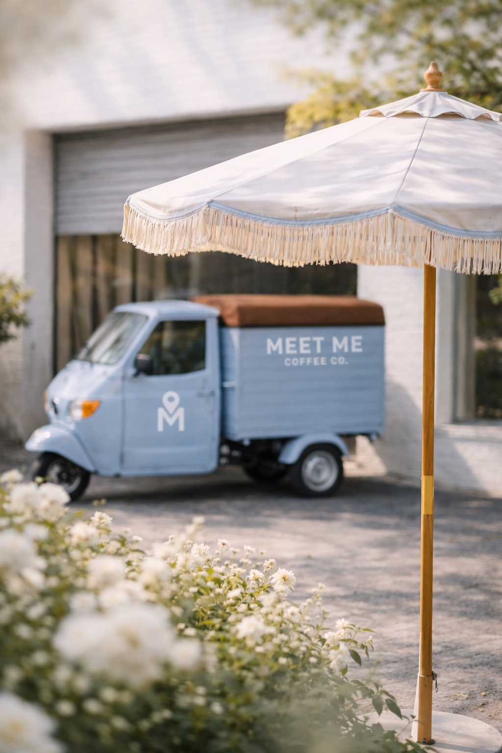 A light-colored umbrella with fringes partially covers a small blue three-wheeled delivery vehicle labeled 'Meet Me Coffee Co.' parked outside a building.