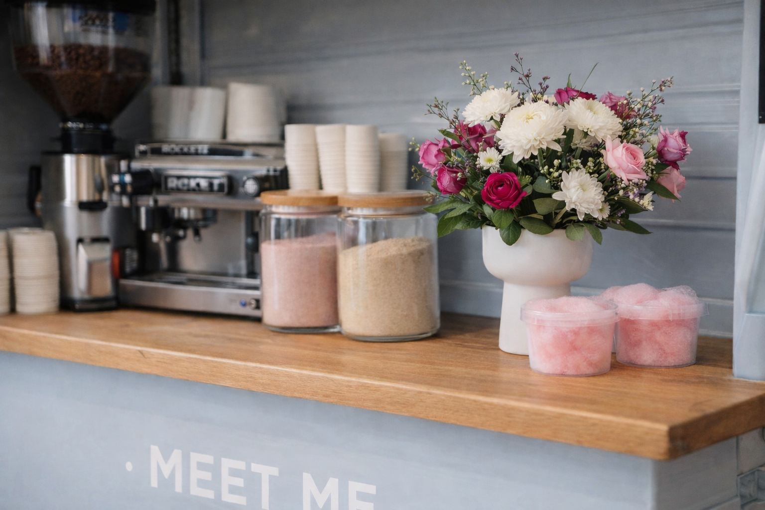 Coffee station with a flower vase and containers of pink and beige powders on a wooden countertop.