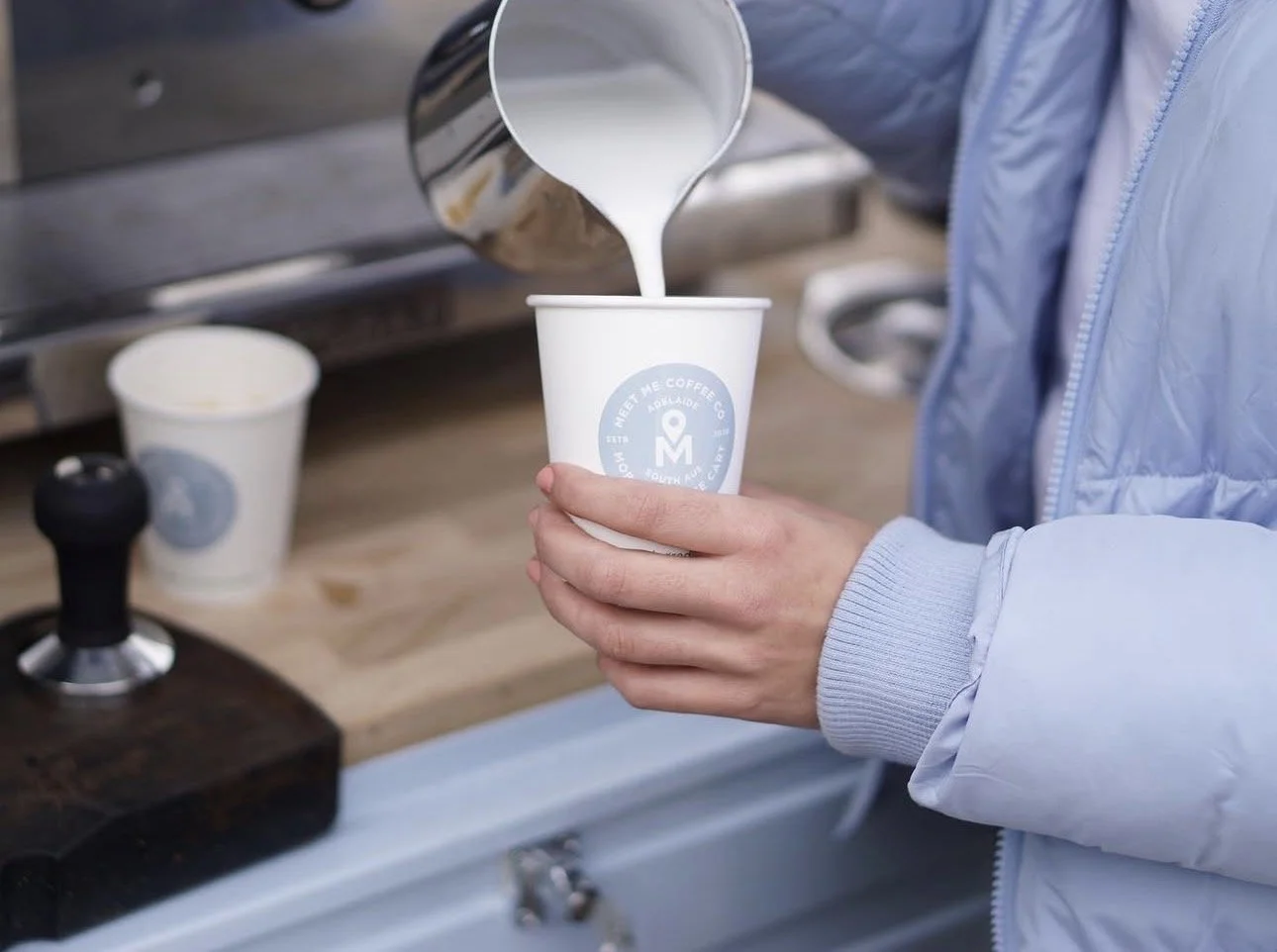 Person pouring milk into a white coffee cup with a logo, on an outdoor wooden counter, with another coffee cup and a tamper nearby.