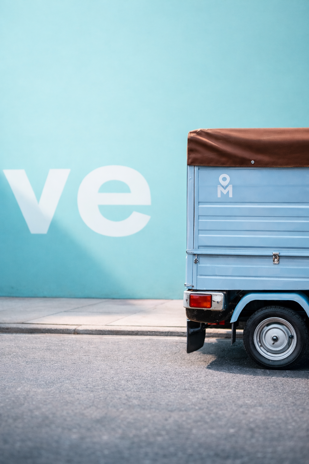 Part of a three-wheeled delivery vehicle with a grey cargo box and a brown cover, parked on a street with a large blue wall featuring white text in the background.