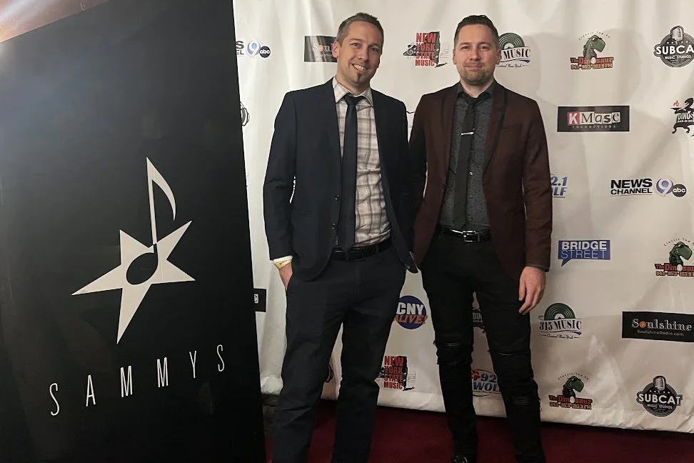 Patrick Blakley and Chris Blakley in suits standing on a red carpet in front of a step and repeat banner with various logos, including Sammys, and a black sign with a star and a musical note.