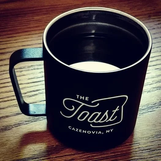 Black coffee mug with the logo 'The Toast, Cazenovia, NY' on a wooden table.