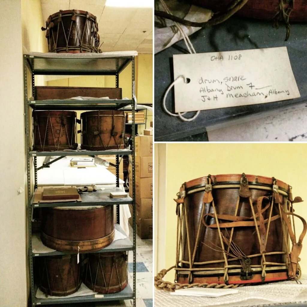 A collection of vintage drums, including a large bass drum, with a smaller snare drum on a metal storage rack. There is a close-up of a snare drum with a handwritten tag that says 'drum, snare, Albany Drum 7, JH Meacham, Albany.'