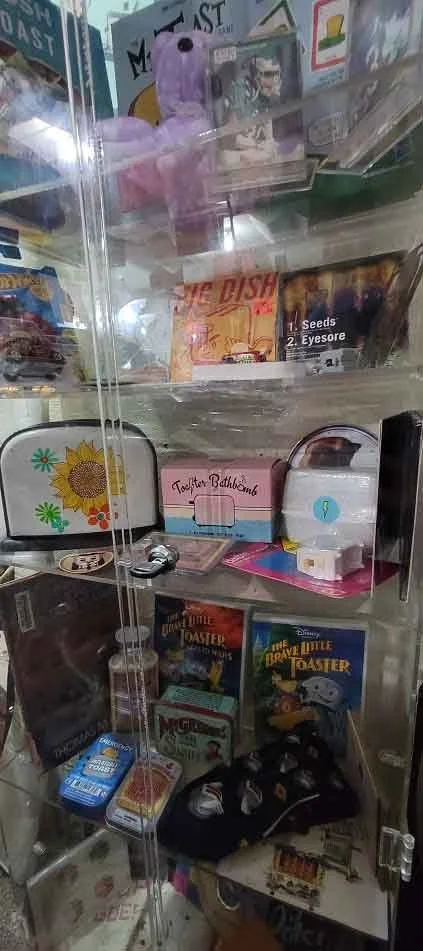 A glass display case containing various toys, books, and small items such as a purple stuffed animal, colorful tin, bath bomb box, and boxed games.