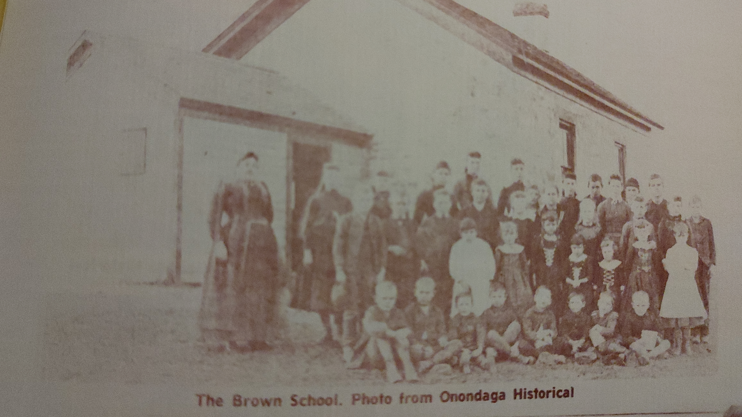 A sepia-toned historical photo of a large group of children and adults standing and sitting in front of a school building, labeled 'The Brown School. Photo from Onondaga Historical' at the bottom.