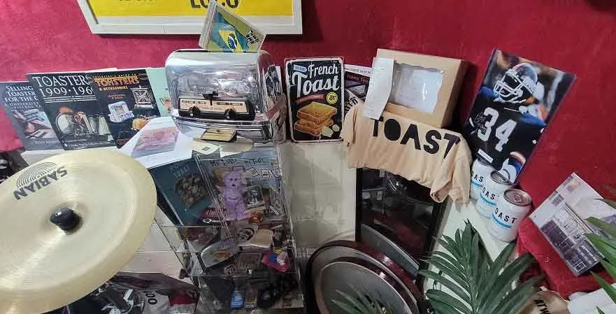 A cluttered tabletop with various items including a drum cymbal, a clear plastic display case, a cardboard box labeled 'French Toast,' a paper bag with 'TOAST' printed on it, a cardboard cutout with the number 34, and several magazine or brochure pag