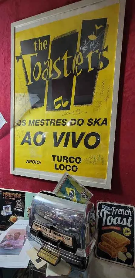 A poster advertising a live performance by The Toasters, a ska band, with support from Turco Loco, mounted on a wall behind a vintage paper money and coin changer, and a French toast advertisement on a table.